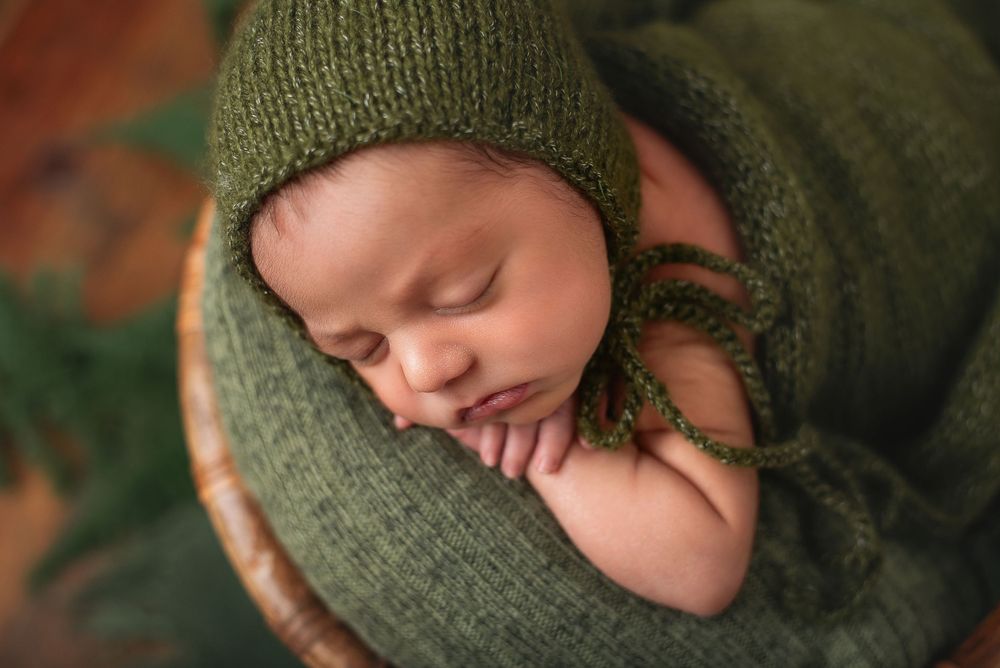 Sleeping newborn wearing olive green knit hat and wrap in rustic setting.