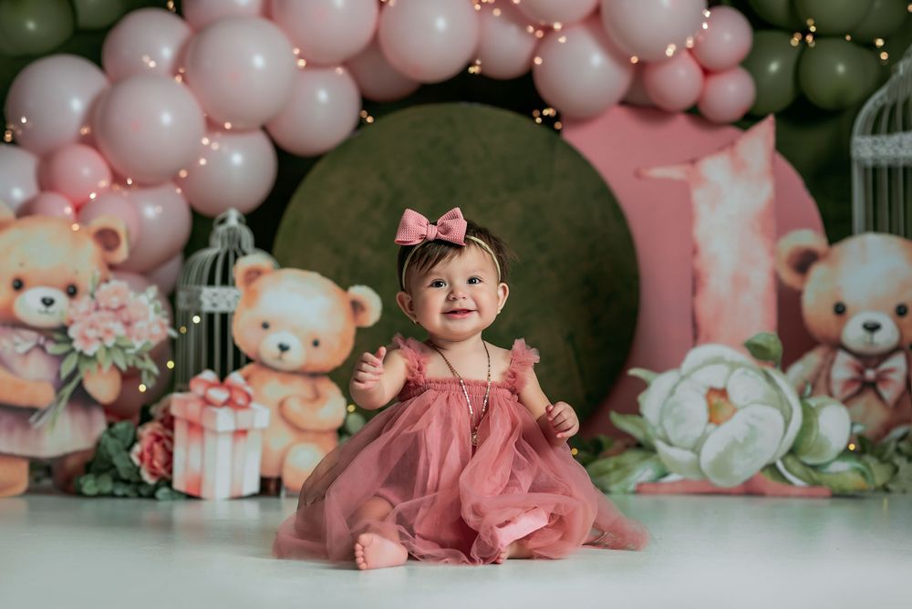 Baby in pink dress sitting among balloon arch and teddy bear decorations.