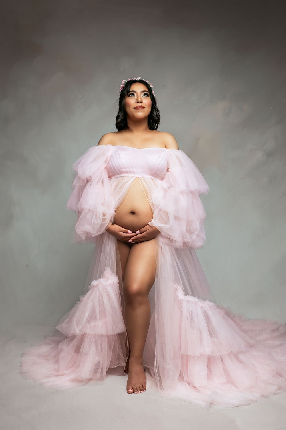 Maternity photo featuring a flowing pink tulle robe against a grey backdrop in glamour style.