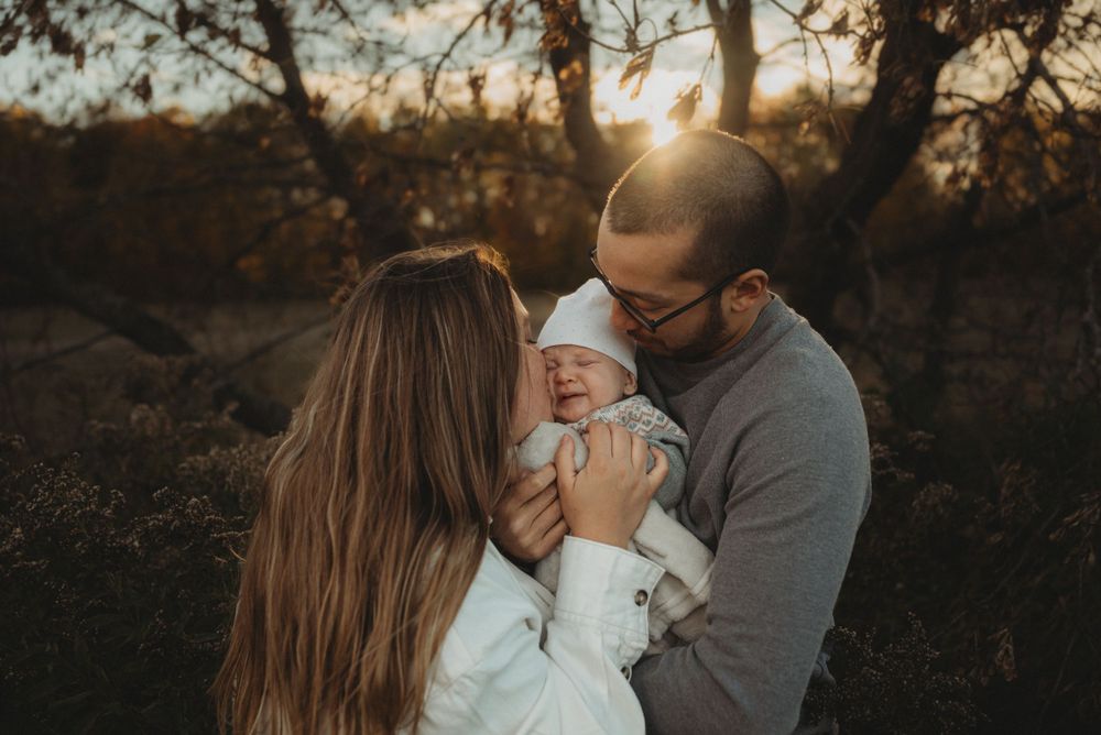 A loving family moment at sunset as they embrace their infant child in a wooded outdoor setting.