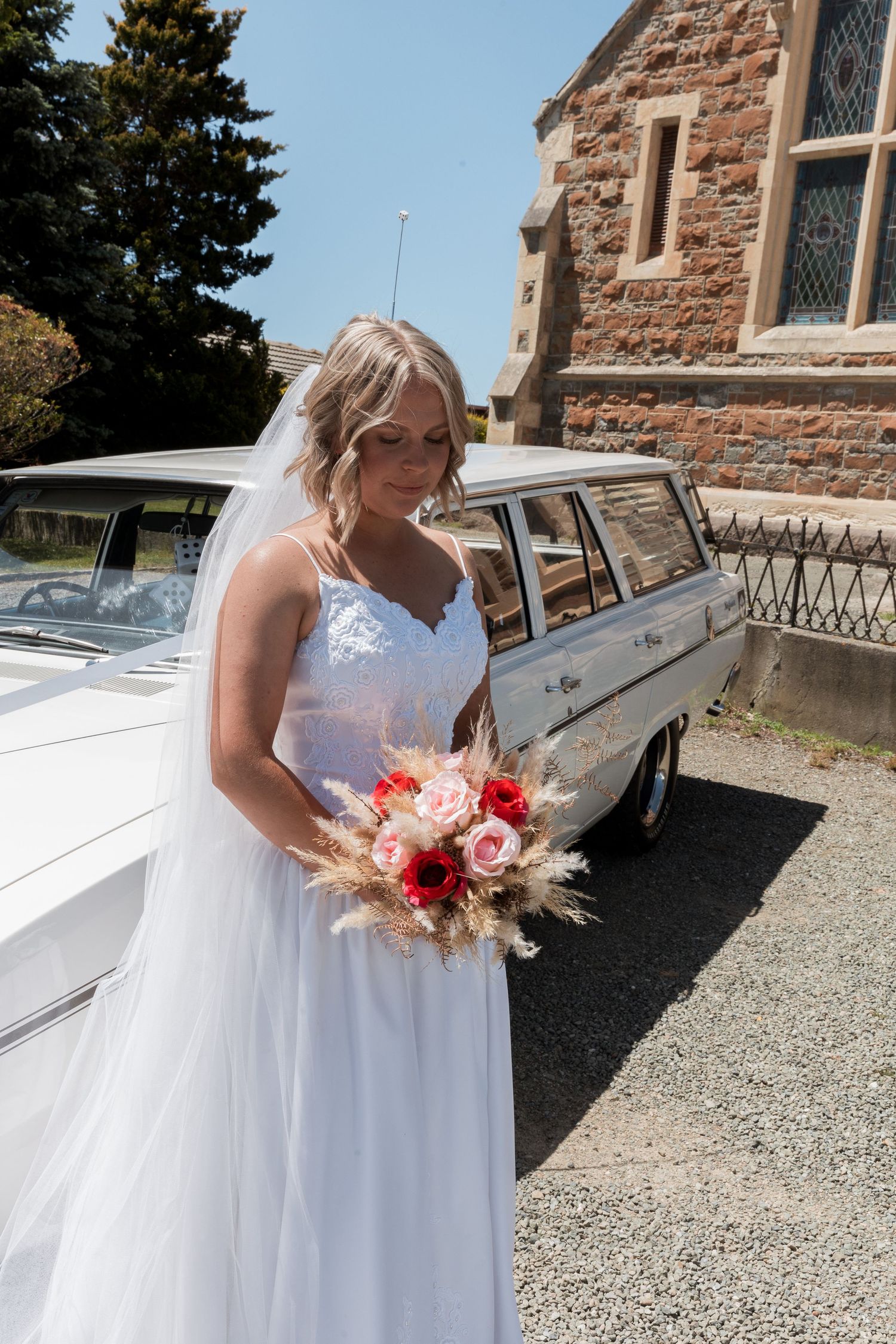 Beautiful wedding attire with flowing white dress and colorful bouquet beside vintage car.
