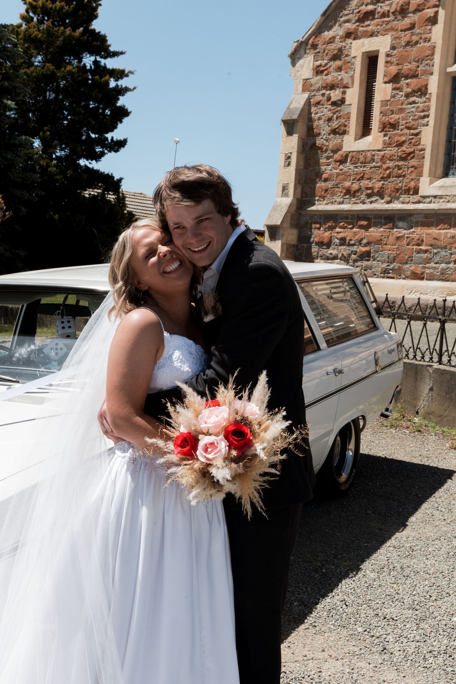 Joyful wedding moment captured beside vintage white car at stone church.