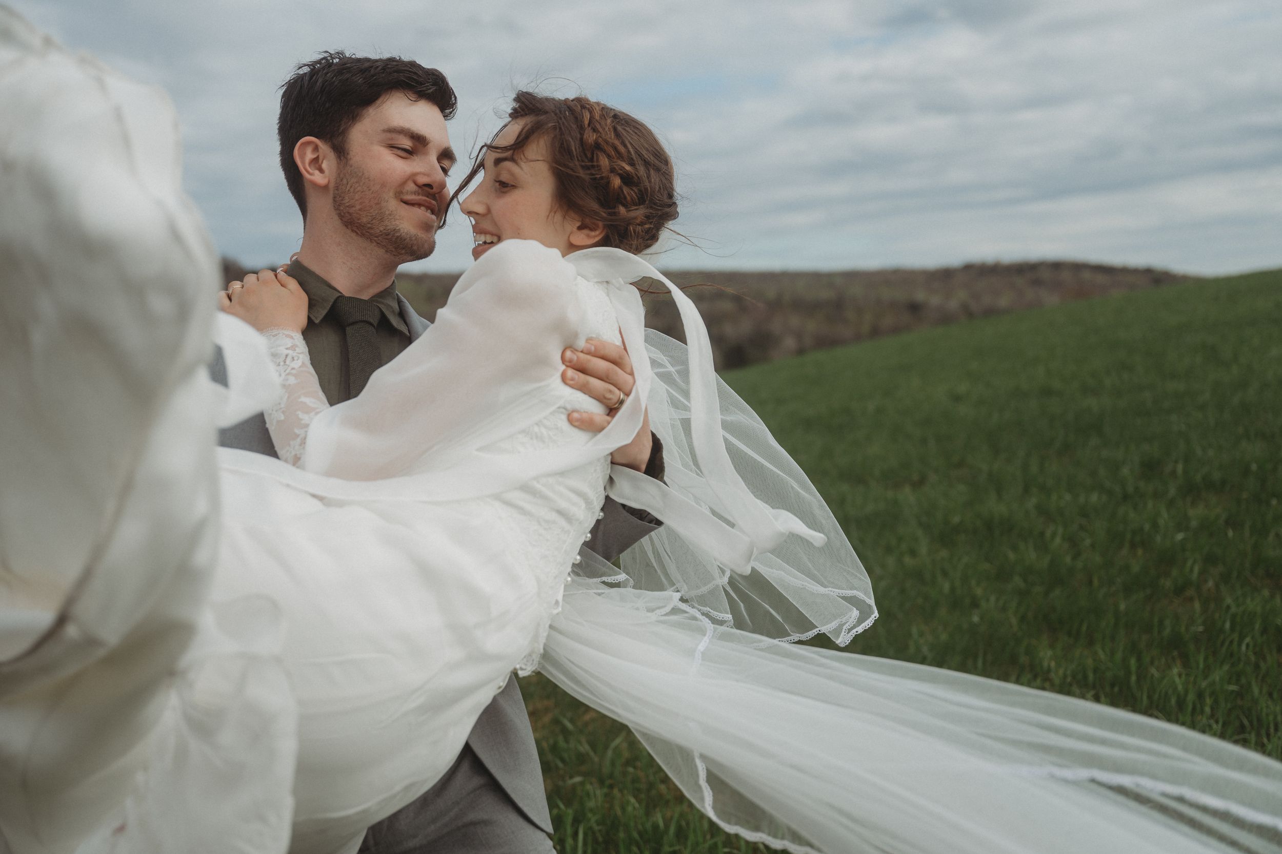 Romantic embrace in white dress and grey suit against green countryside landscape.