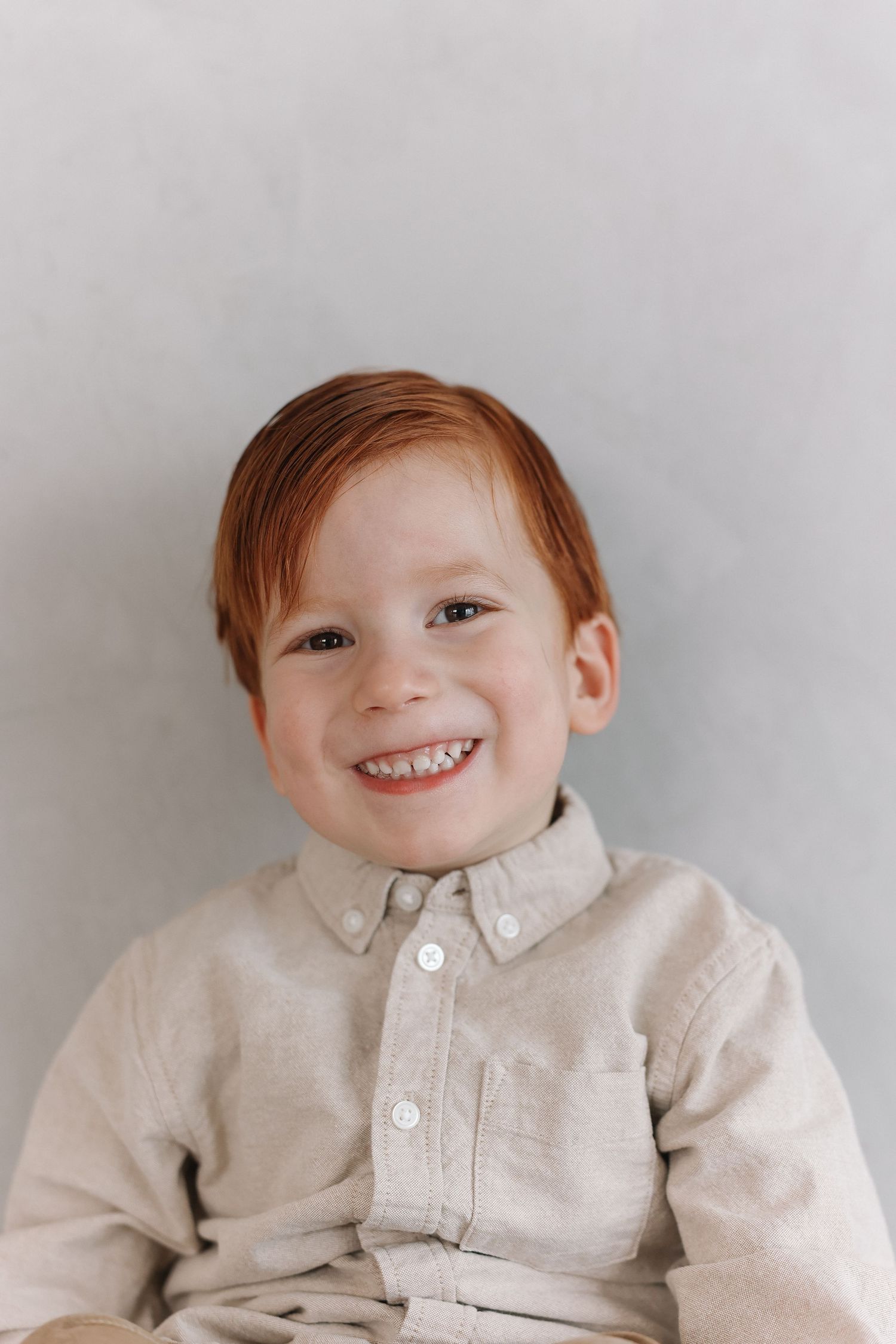 Young child with red hair wearing a beige button-down shirt smiles while posing against a light gray background.