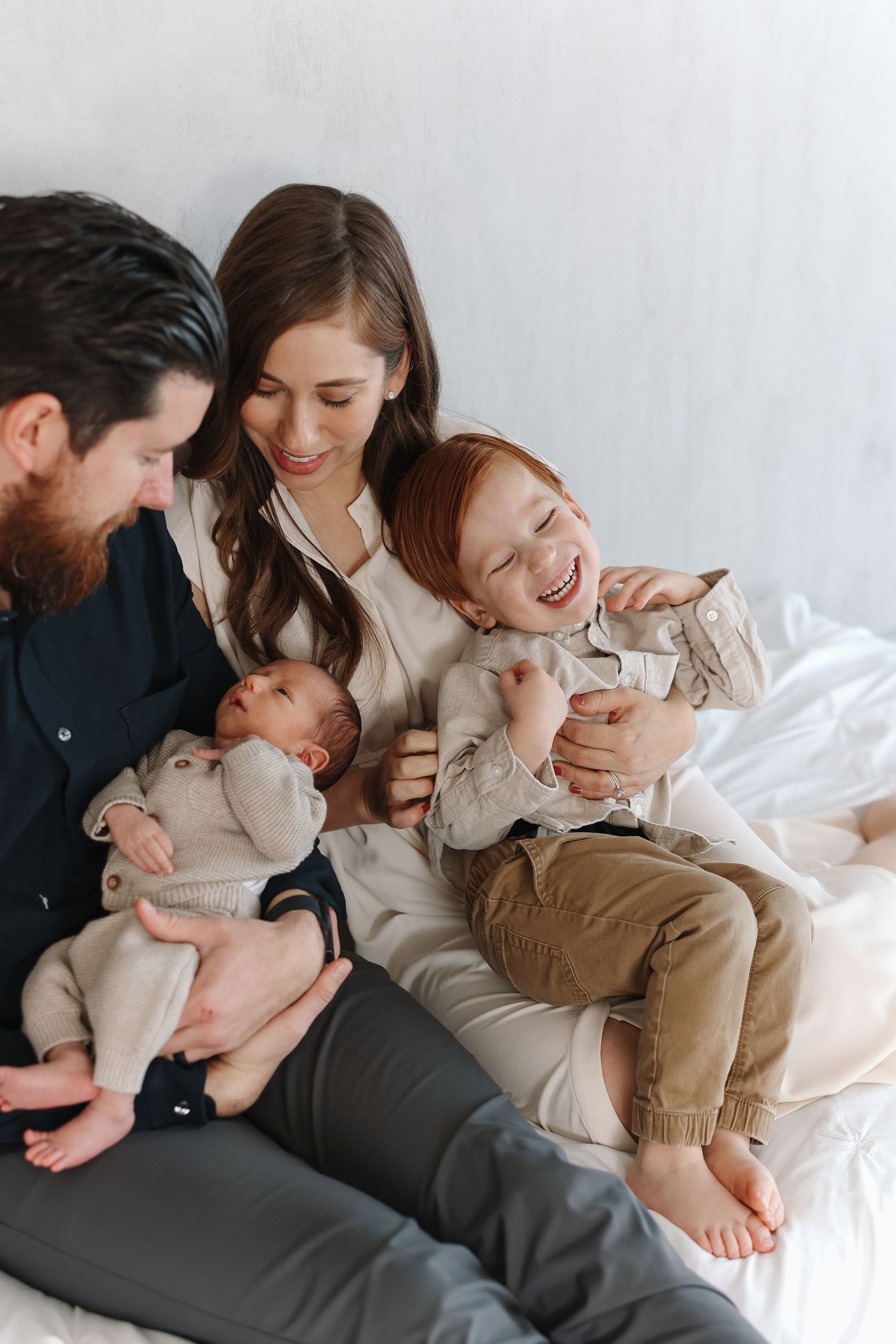 Parents cuddle and bond with newborn baby while lying comfortably on white bedding together.