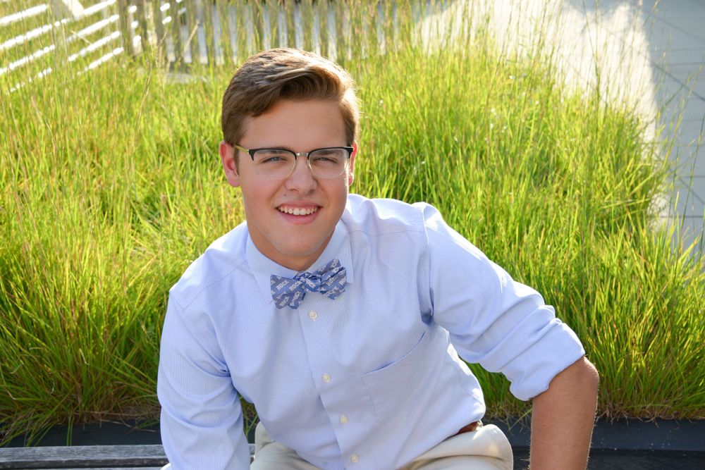 Young person in bow tie and white shirt sitting outdoors by tall grass.