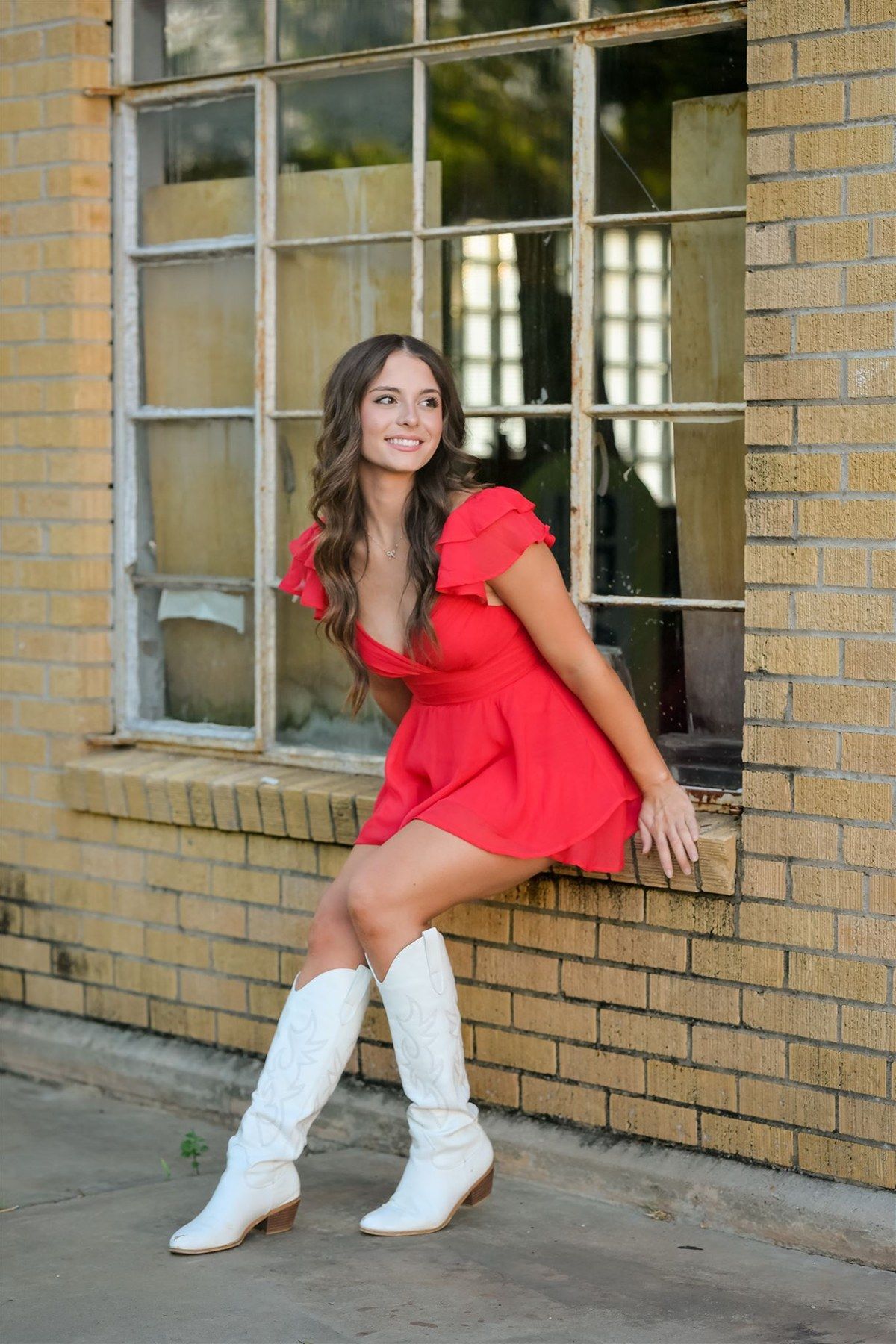 A person in a red dress and white knee-high boots poses by a brick wall with windowpanes.