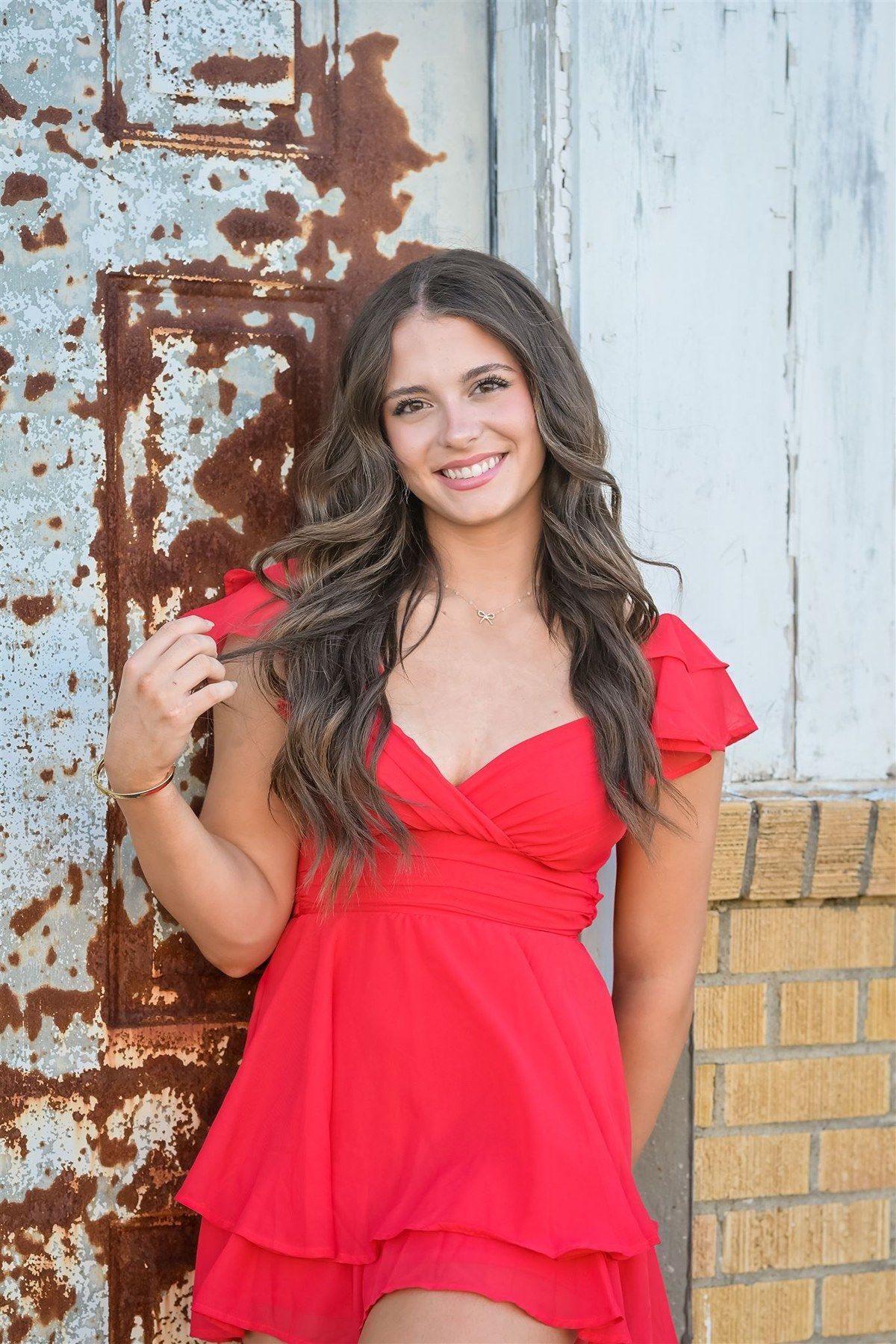 Woman with long wavy brown hair poses against rustic door wearing a red summer dress with cap sleeves.