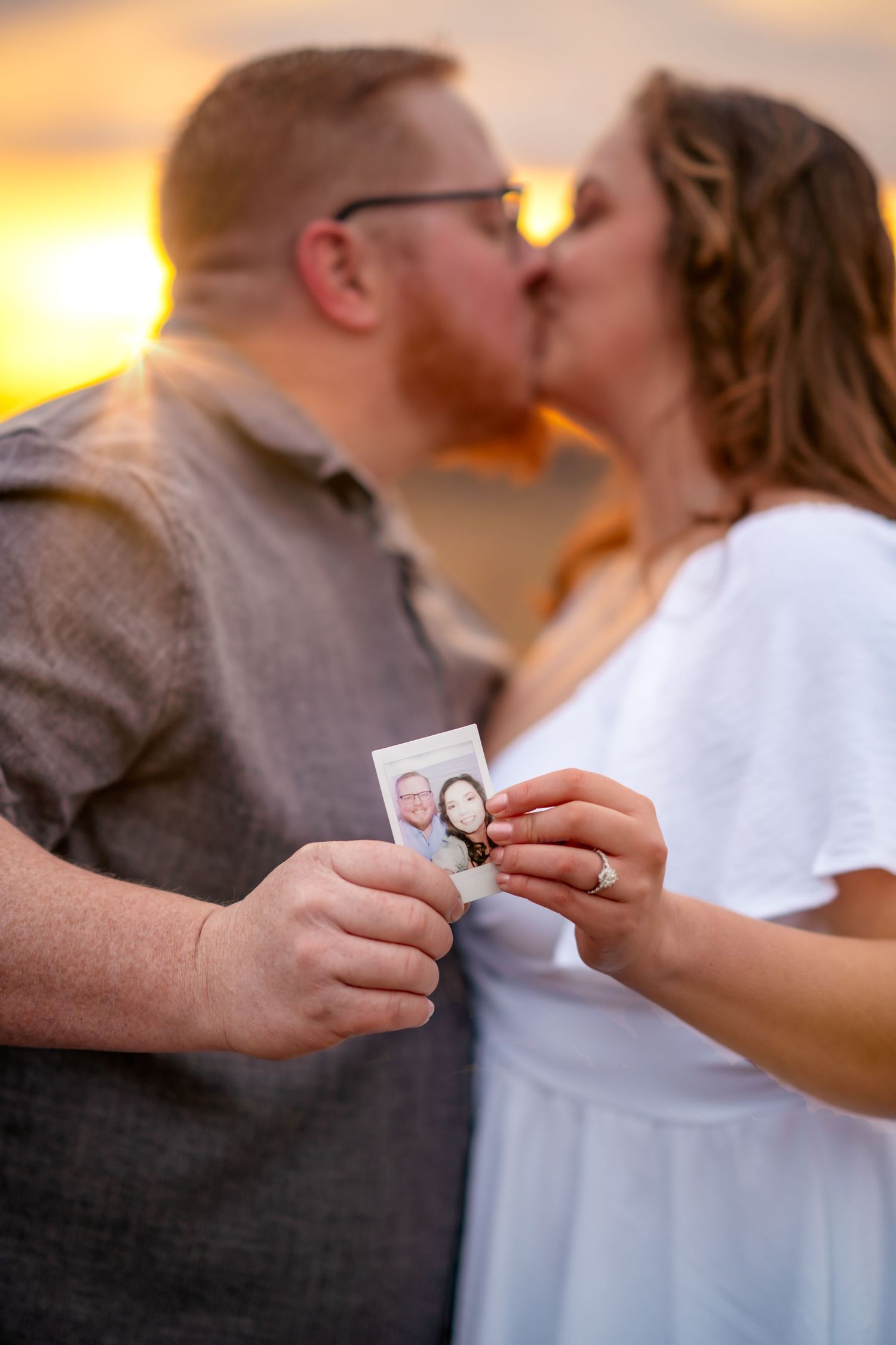 A couple kisses at sunset while holding an engagement ring, captured in a romantic moment.