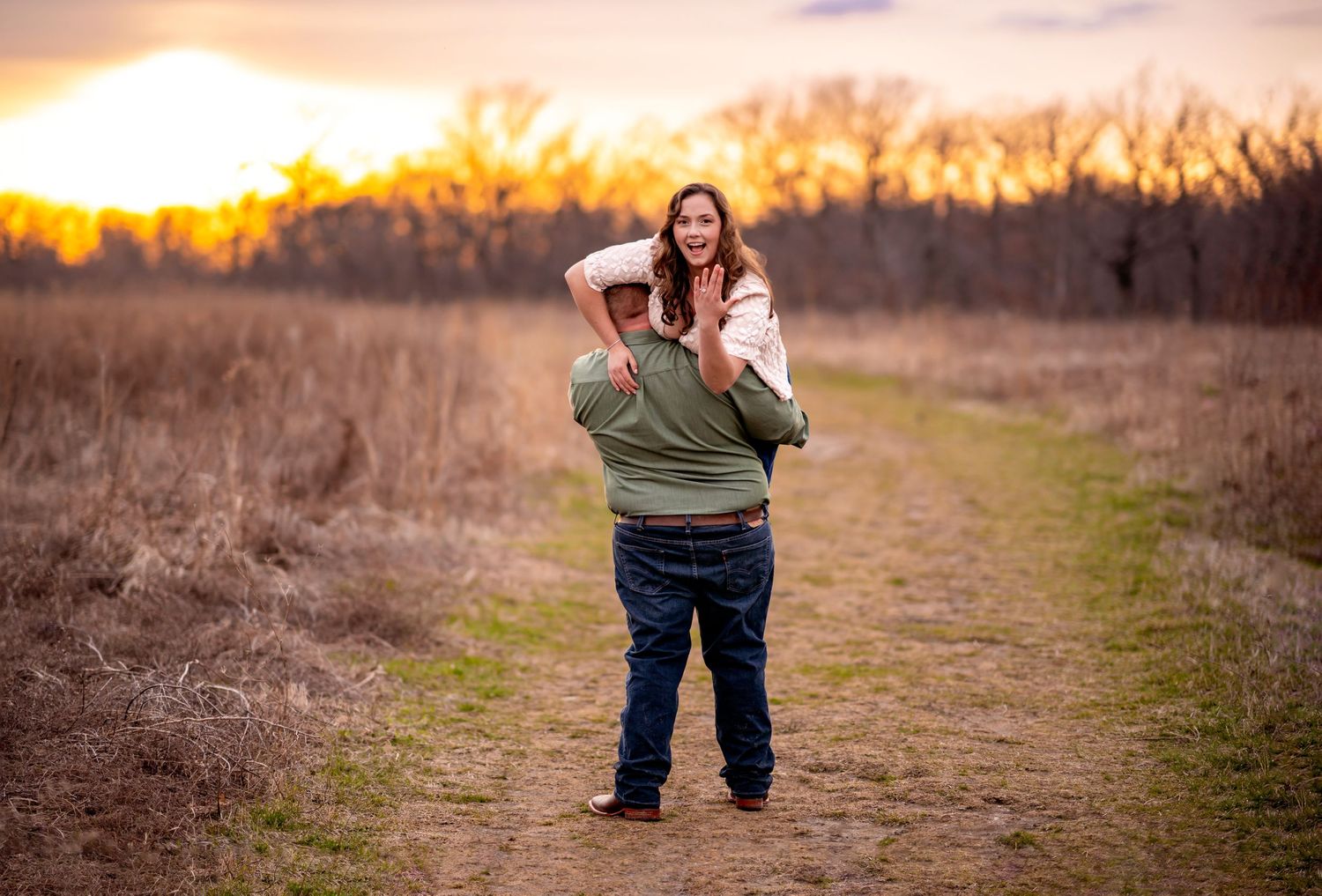 A couple shares a romantic moment on a dirt path during golden hour sunset with dried grass in the background.