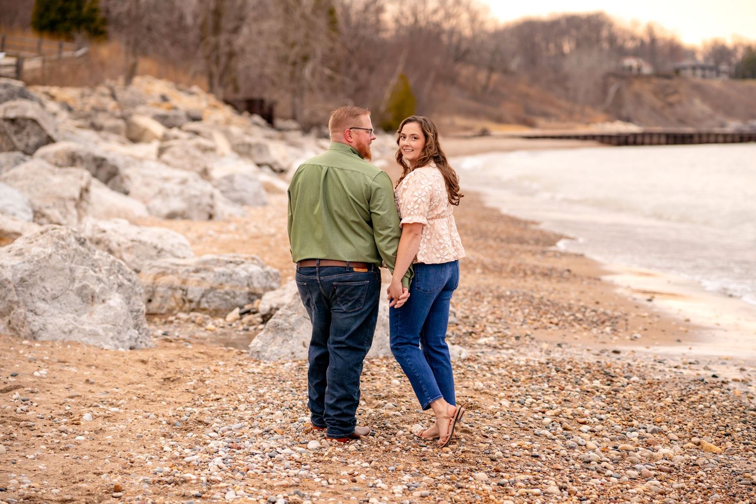 A couple embraces while standing on a rocky shoreline at sunset with warm lighting.