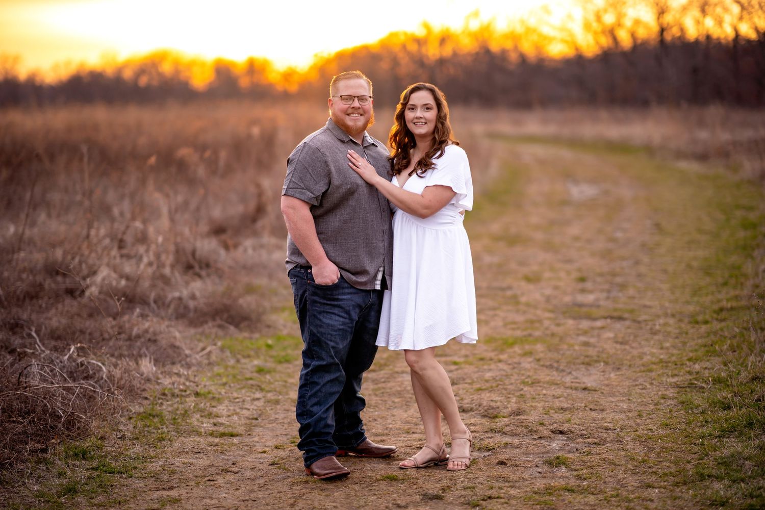 A couple poses together outdoors on a dirt path during golden hour sunset with glowing golden light behind them.
