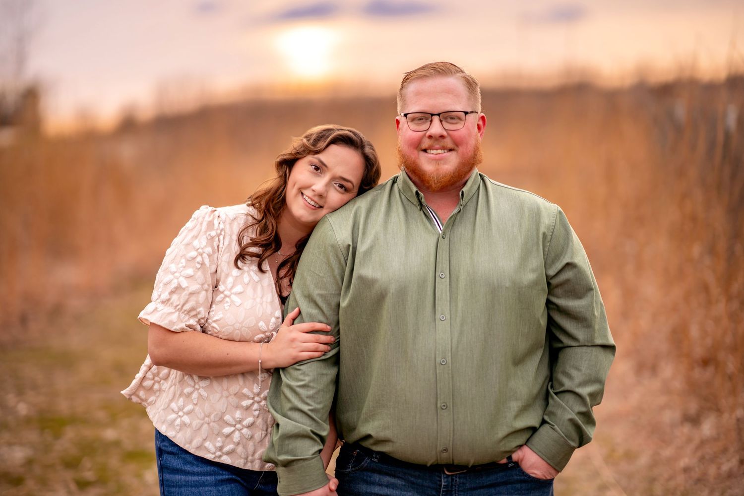 A couple embraces in a golden field at sunset with tall grass and warm lighting in the background.