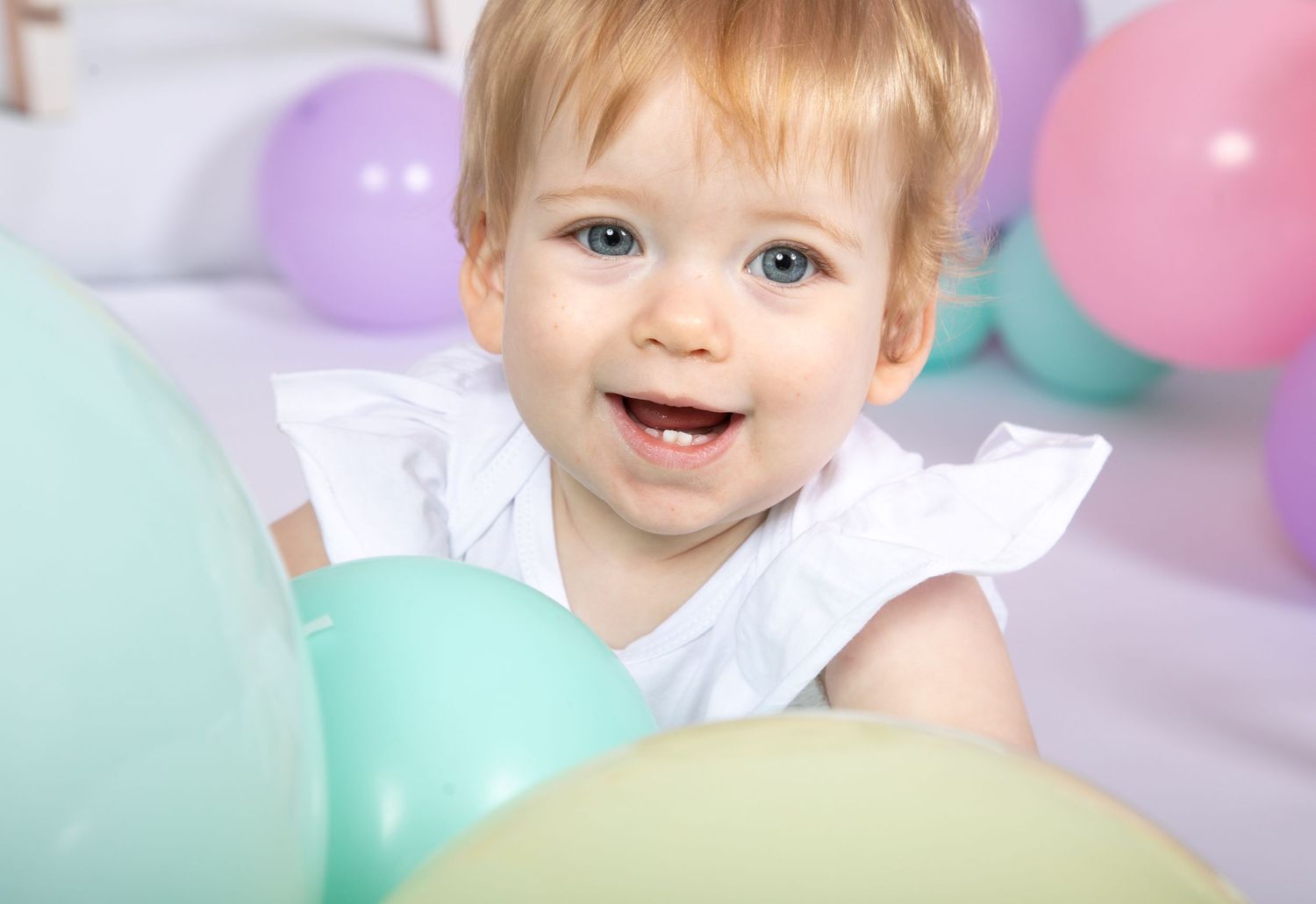 A young child smiles happily surrounded by pastel balloons at a party celebration.