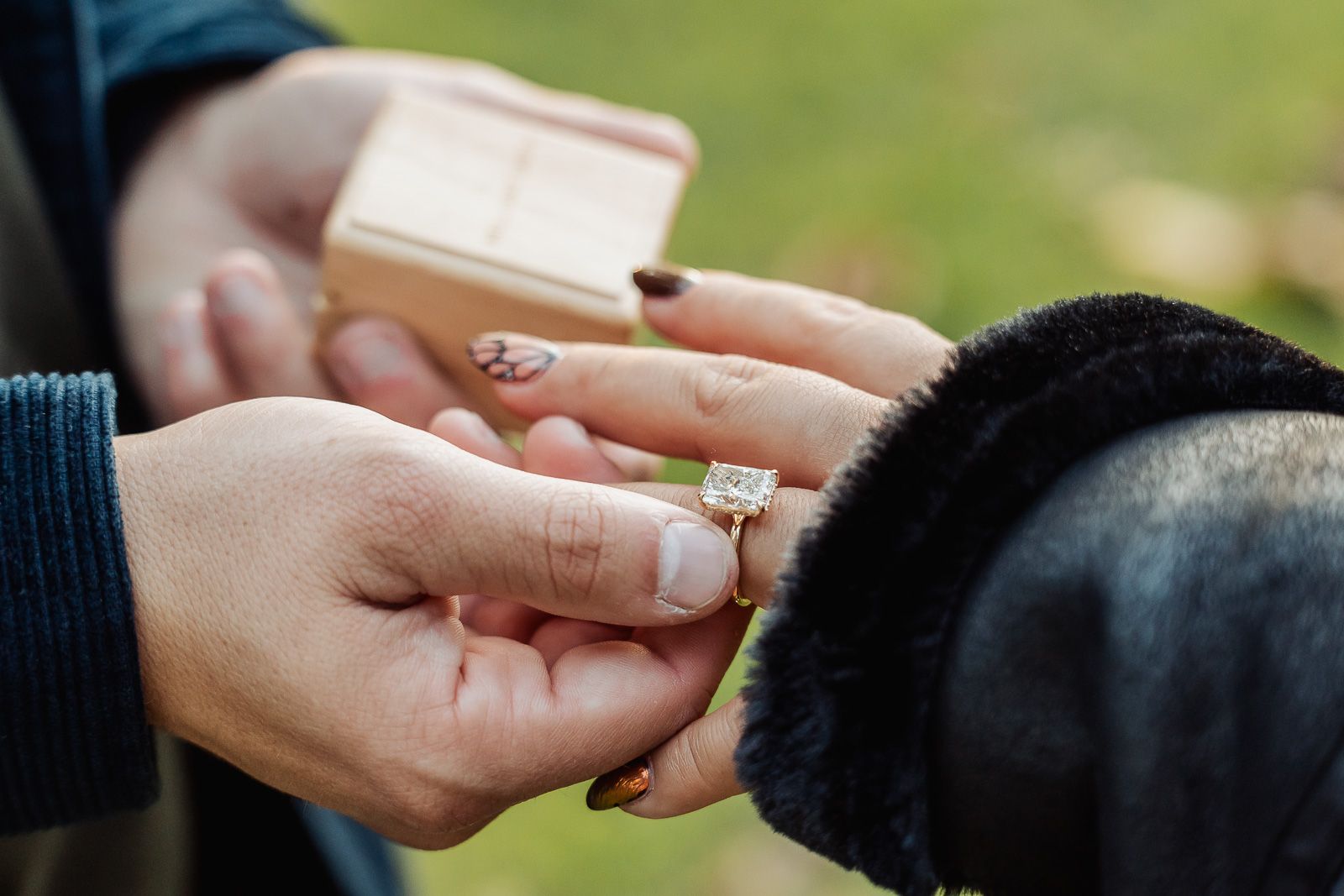 Close-up of hands presenting an engagement ring in a wooden box outdoors.