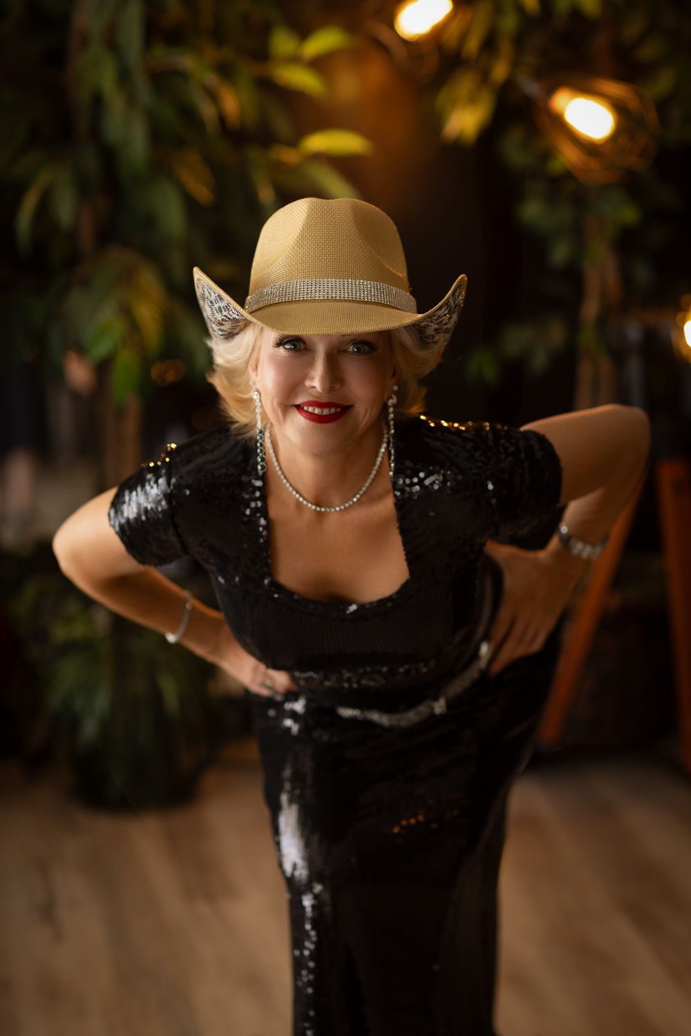 A glamorous figure in a sparkling black evening gown and beige fedora poses dramatically against a dark background.