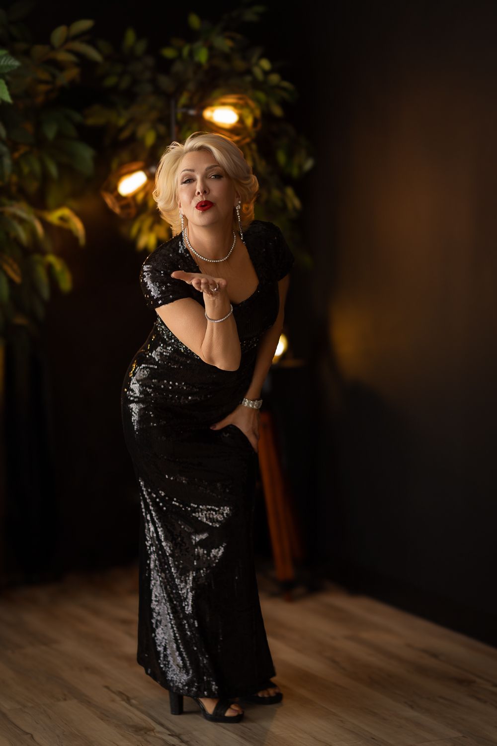 A person strikes a glamorous pose in a sparkling black sequin evening gown on a wooden floor with ambient lighting.