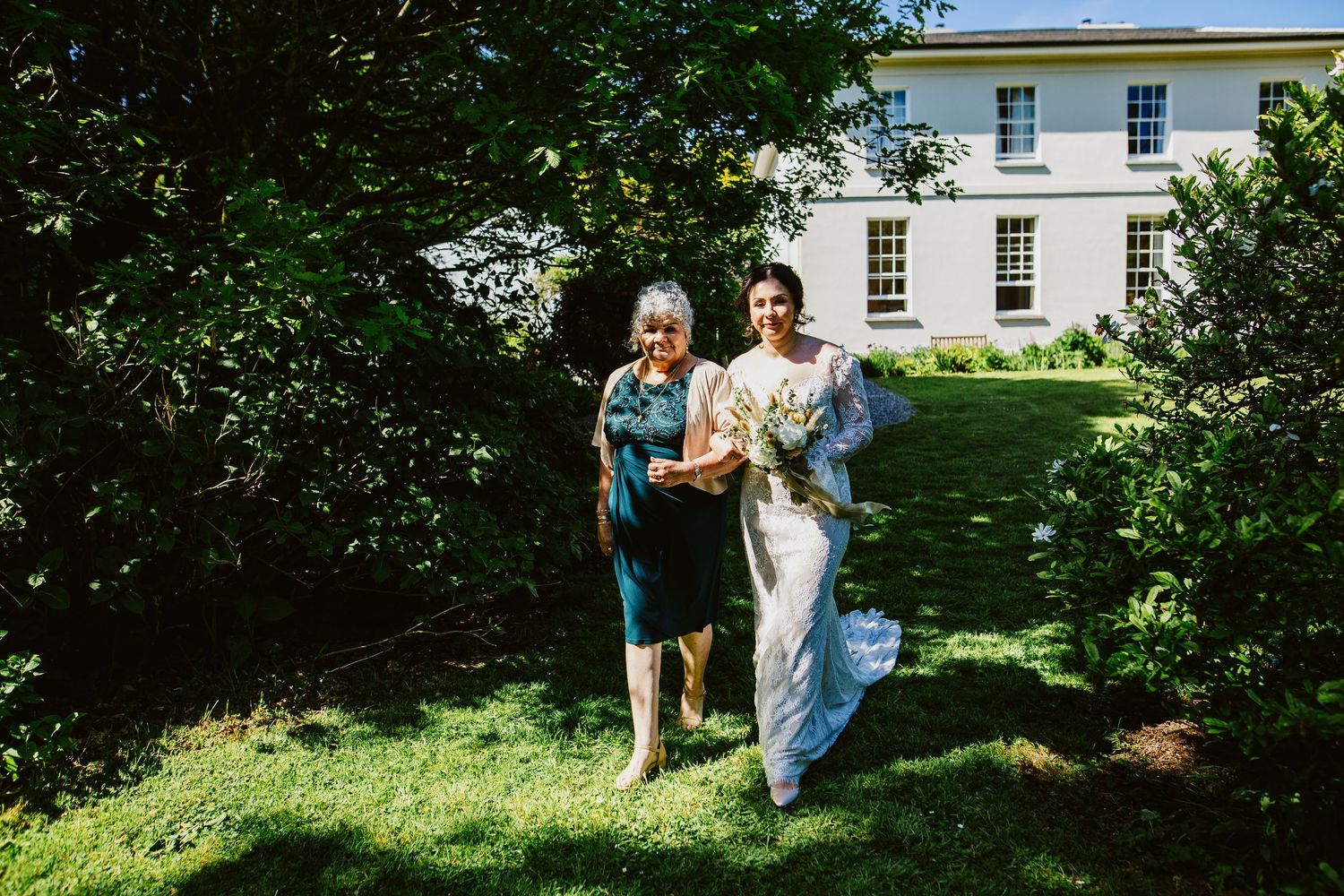 Two people walking together on a lush green lawn with a white manor house in the background.