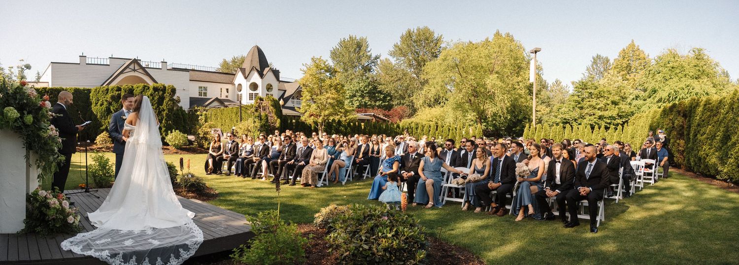 Panoramic view of an outdoor wedding ceremony with guests seated on white chairs under a floral arch at golden hour.