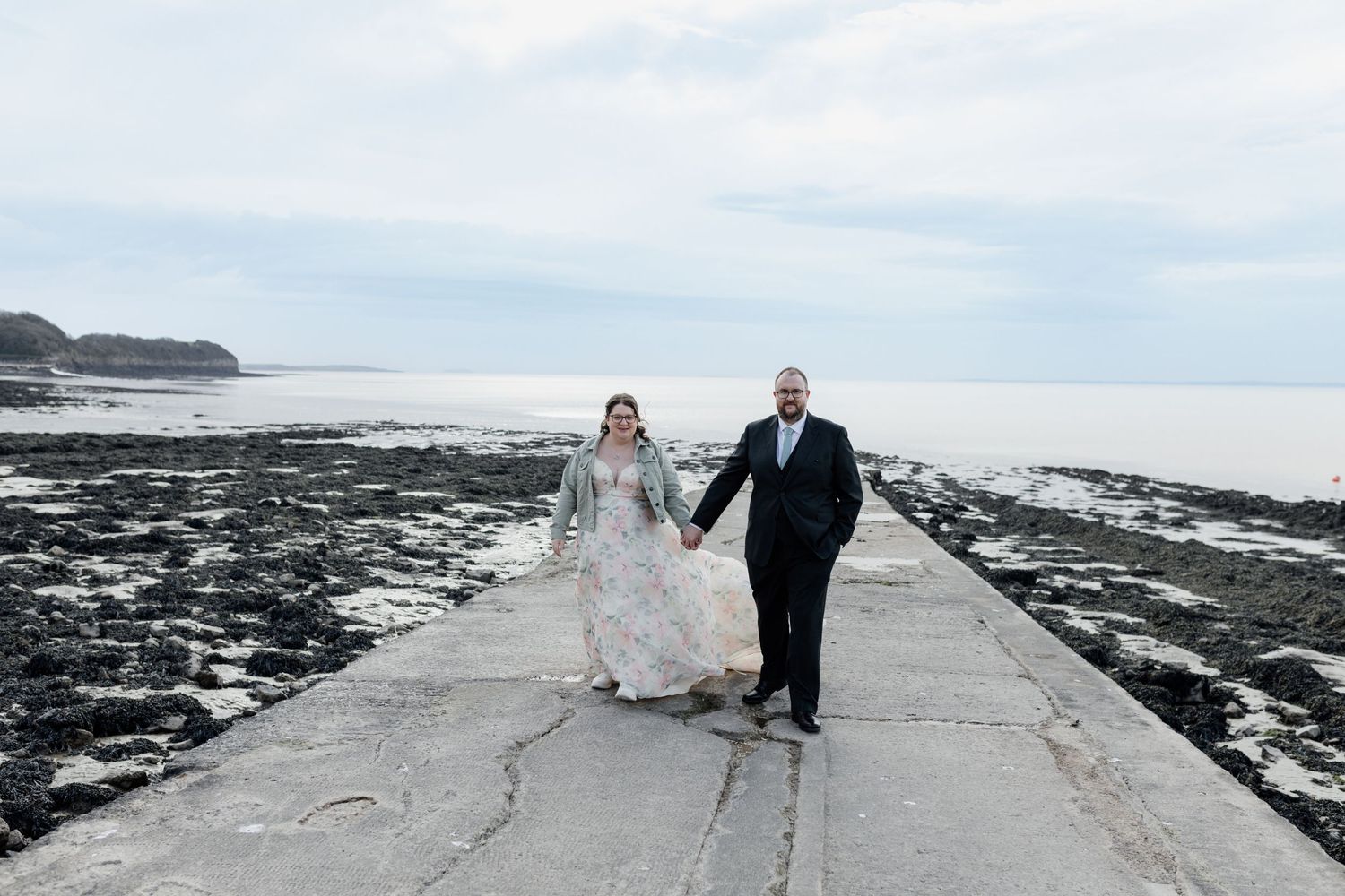 A couple in wedding attire walks along a rocky coastal path with dramatic ocean views and cloudy skies in the background.
