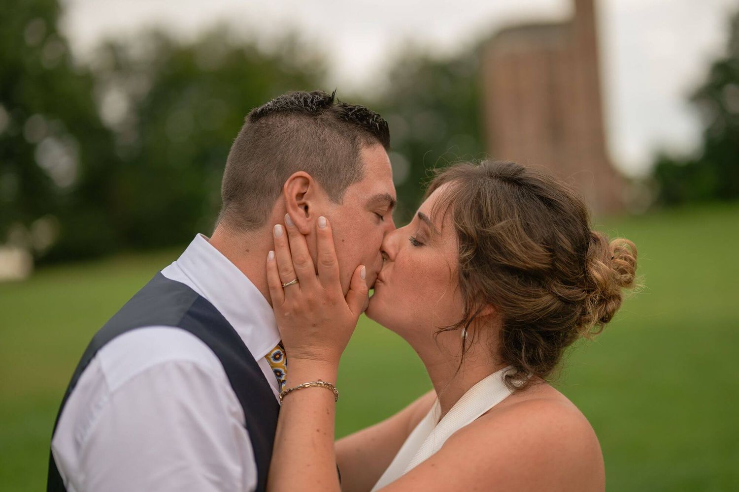 A romantic couple shares a tender kiss outdoors in a lush green setting during their wedding celebration.