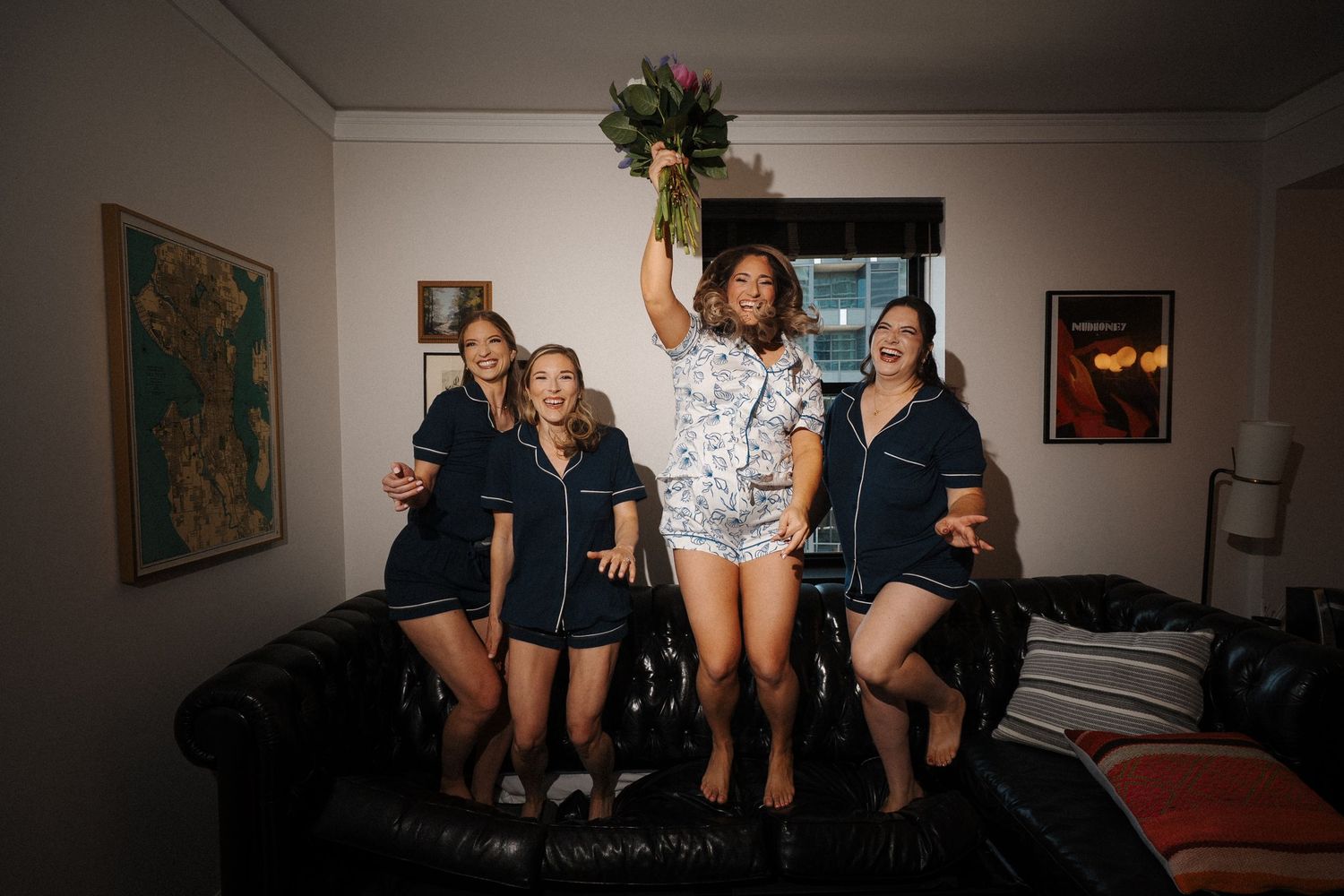 A group of energetic people jumping and posing on a black leather couch in a living room setting.