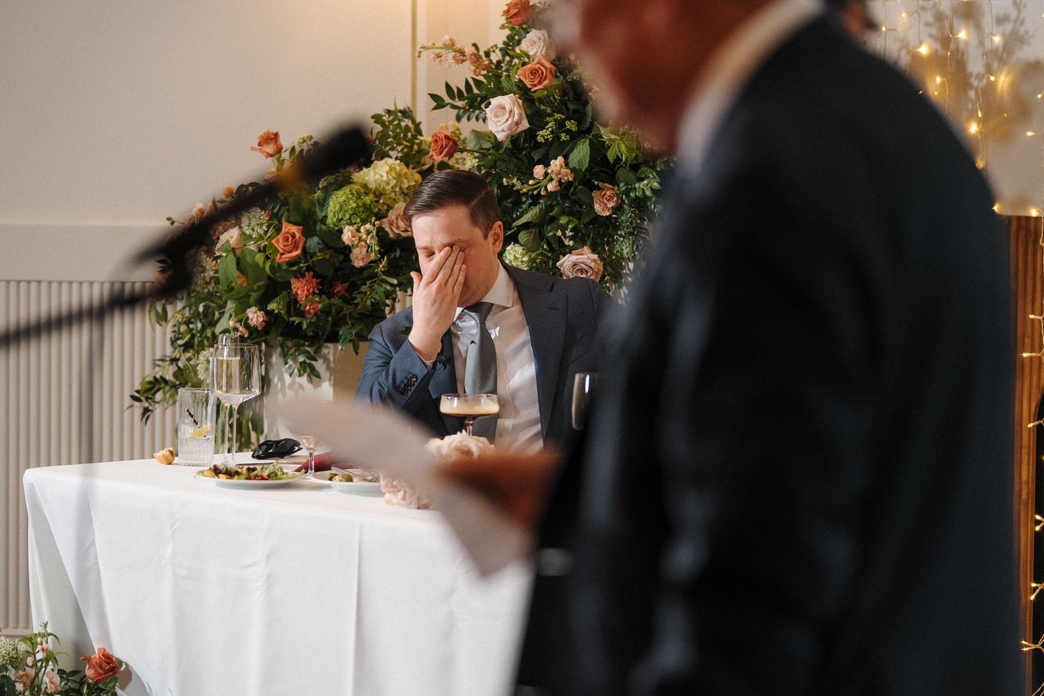 Someone in a suit shows an emotional reaction during a wedding reception with floral decor and white tablecloths.
