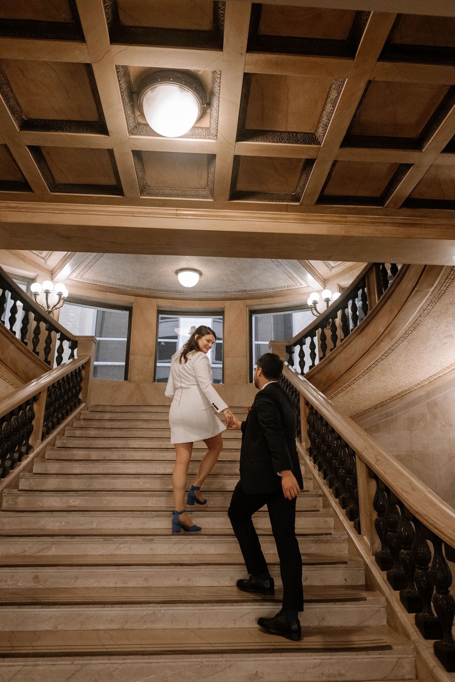 Two figures ascend a grand interior staircase with ornate wooden railings and warm lighting from above.