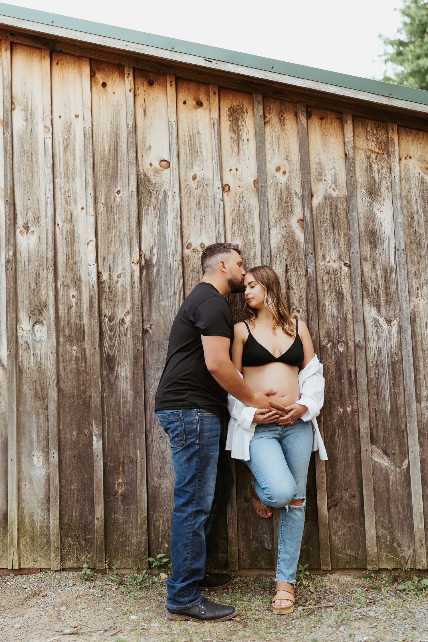 maternity photos in pittsburgh pennsylvania, man and woman pose against a brown barn, his hand on her pregnant belly, kissing her forehead