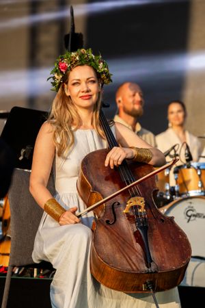 Musician in white dress and floral crown performs with cello on outdoor stage.