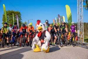 Mascot in rooster costume posing with cyclists at starting line of race.