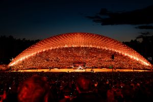 Illuminated amphitheater glowing red at night with crowd silhouettes and dramatic arched roof structure.