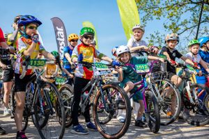 Group of cyclists gathered at starting line with colorful flags and banners.