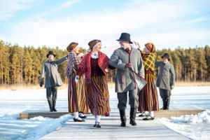 People in traditional folk costumes dancing on snowy wooden pier in winter landscape.