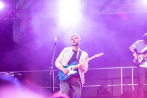 Concert performer playing guitar on stage bathed in purple spotlight.