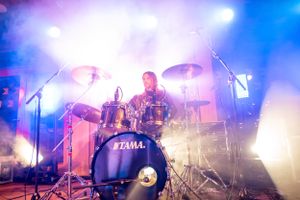 Drummer performs energetically behind Tama drum kit with colorful stage lighting.