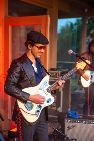 Guitarist in leather jacket and cap plays white electric guitar during performance.