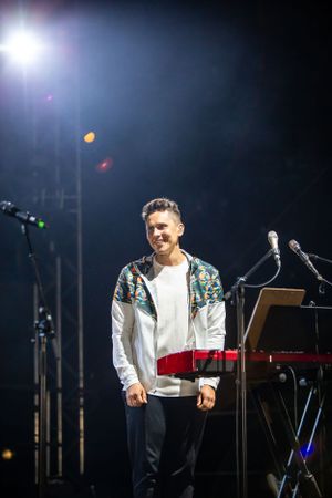 Performer in patterned jacket stands on stage with keyboard setup and stage lighting.