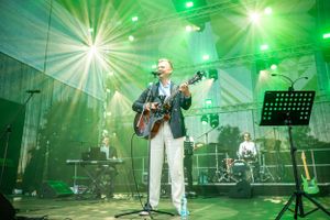 Concert performer playing guitar on stage with green lighting effects and band equipment in background.