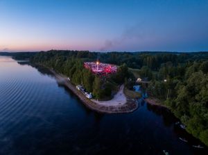 Aerial twilight view of illuminated festival grounds near waterfront surrounded by forest.