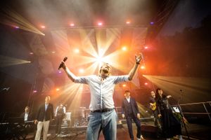 Performer on stage with dramatic lighting raising arms triumphantly during concert performance.