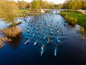 Aerial view of numerous paddleboarders scattered across a scenic lake surrounded by trees.