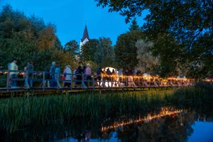 Illuminated outdoor festival at dusk with church spire and crowd reflections in calm water.
