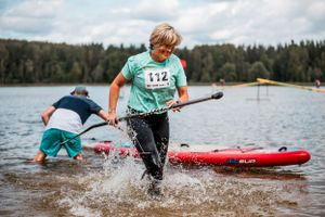 Person in teal shirt splashes through water while running near a paddleboard during a competition.