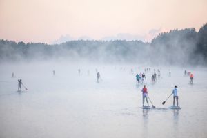 Misty lake scene with multiple paddleboarders silhouetted against foggy mountain backdrop.