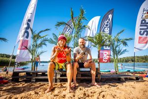 Beachside paddleboarding event with flags and palm trees on a sunny day by the water.