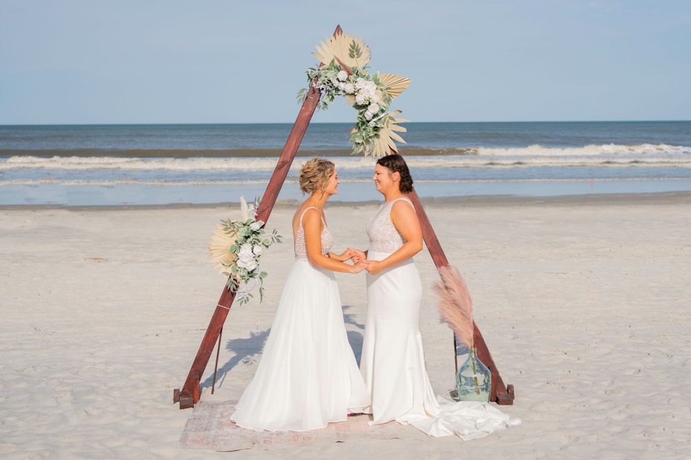 Two brides in white dresses pose under a floral arch on a sunny beach in St. Augustine, Florida