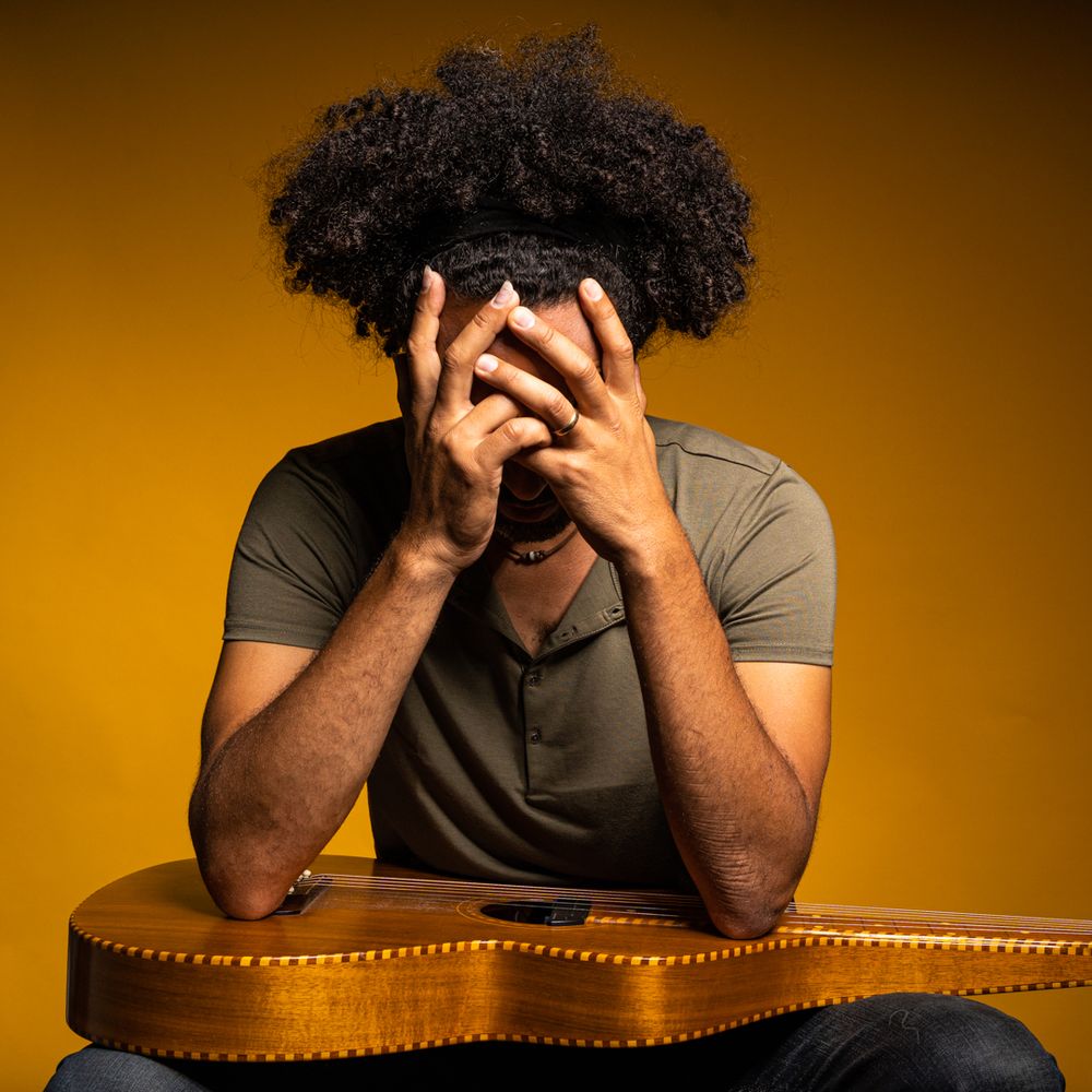 A person with curly hair sitting at a leather chair against a golden brown background holds a camera.