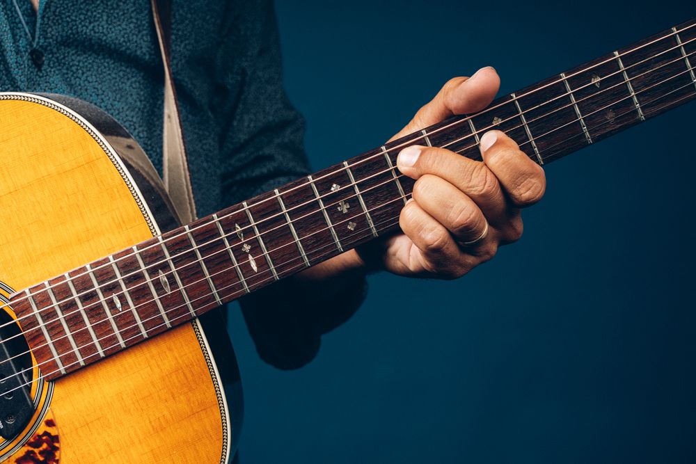 Hands play a chord on a wooden acoustic guitar against a deep teal background.