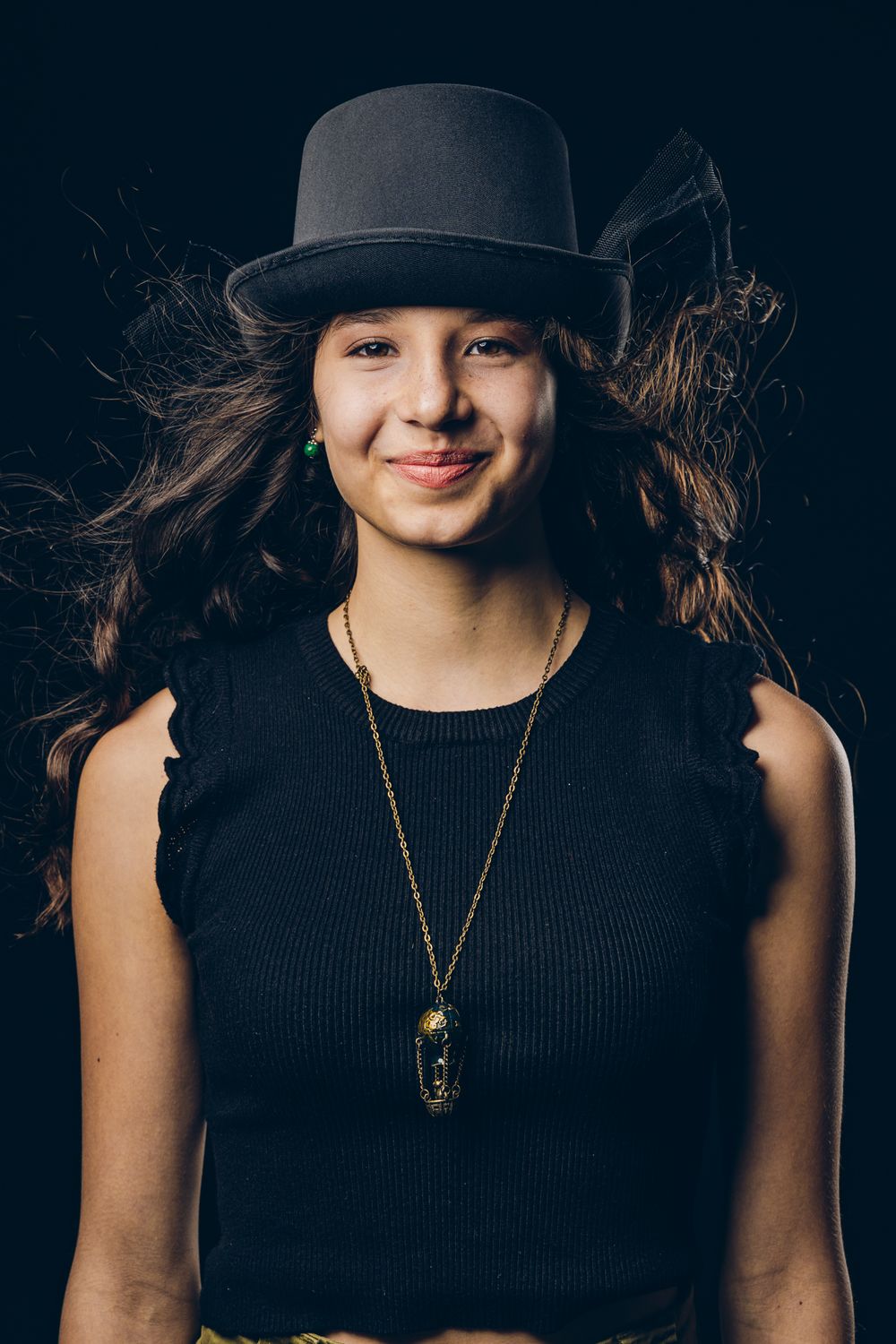 Portrait of a person wearing a black tank top, bowler hat and pendant necklace against a dark background.